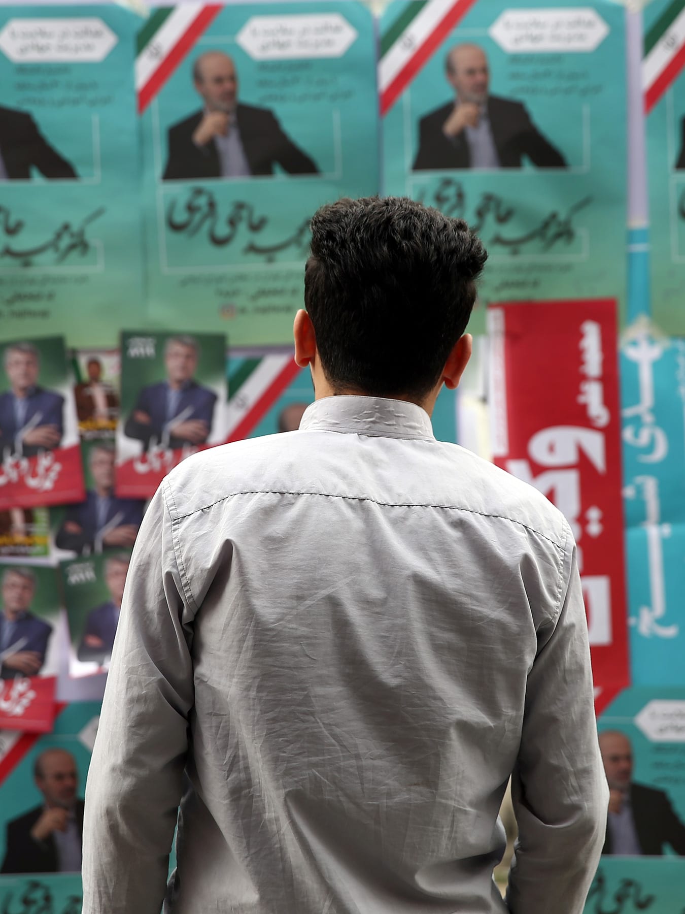 The photo shows the man standing in front of a board that is placarded with multiple political campaign posters, many of them identical and pasted over and over. The predominant colors are green, white, and red—like the Iranian flag. 