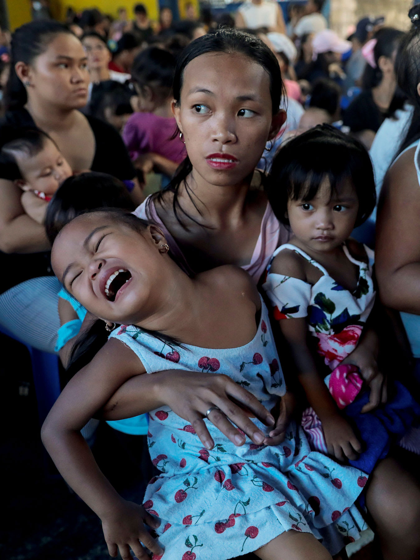 The photo shows a crowd of parents and their children. The woman in the foreground has her arm around her young daughter, who appears to be screaming and struggling to get free.