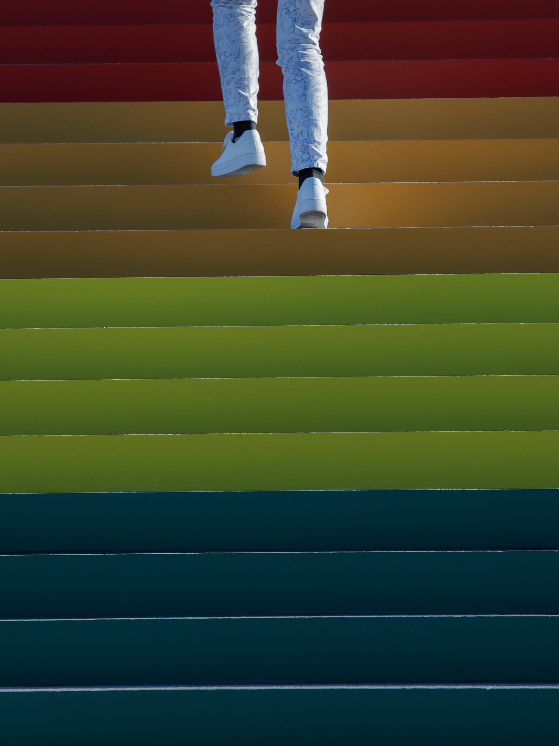 Photo shows the legs and feet of someone wearing white pants and white sneakers walking up a wide staircase that fills the frame and is brightly adorned in rainbow hues. This is a striking photo.