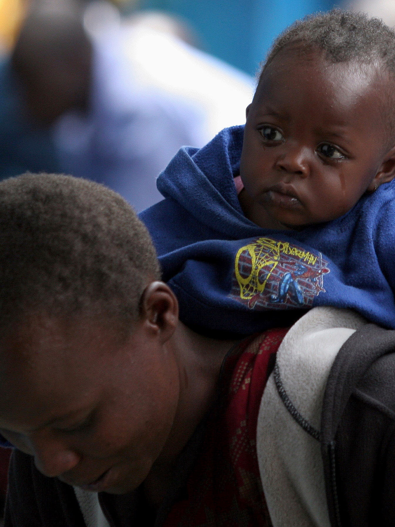 Photo shows a woman leaning down, almost out of frame, with a small child on her back. The child wears a blue spiderman hoodie and has been crying. You can see a tear streak on his cheek. 