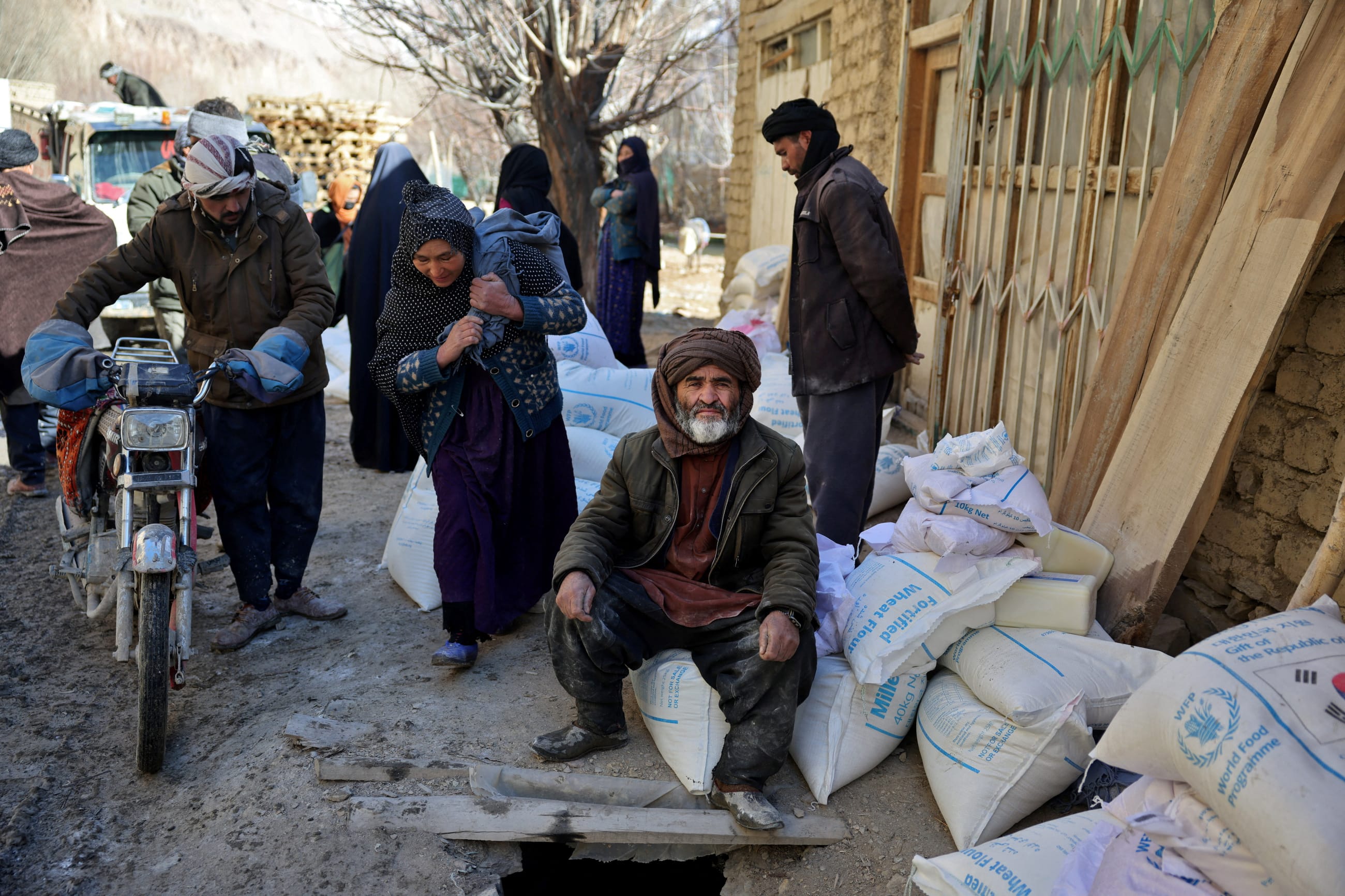 An Afghan man sits on sacks of flour as a woman carries goods on her back after receiving aid at a World Food Program distribution center, in Yakawlang, Bamyan province, Afghanistan, on January 1, 2026.