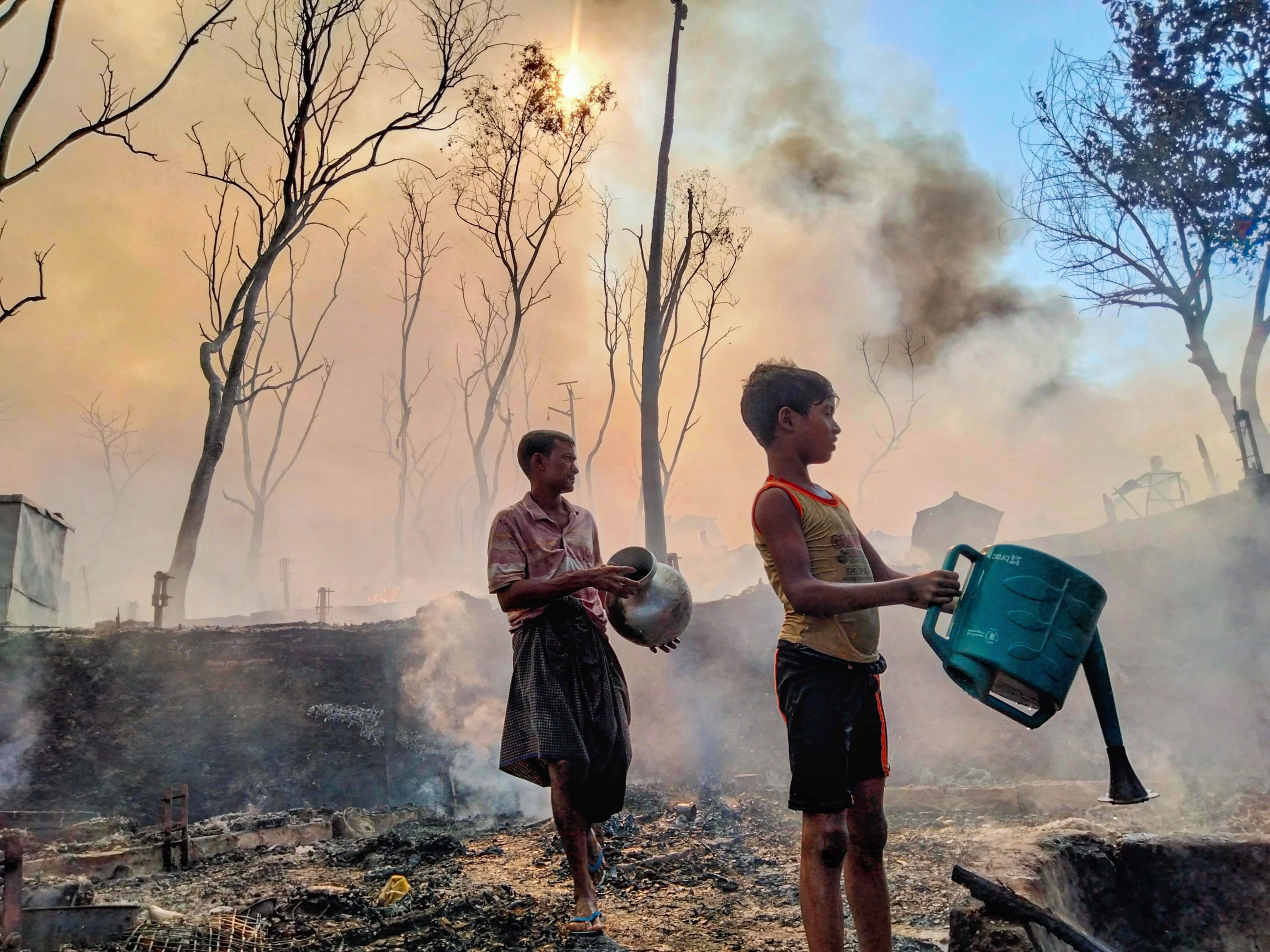 A father and son, working to reclaim what remains of a home reduced to ash, douse smoldering earth with water after a fire tore through their section of the encampment, in Cox's Bazar, Bangladesh, on December 24, 2024.