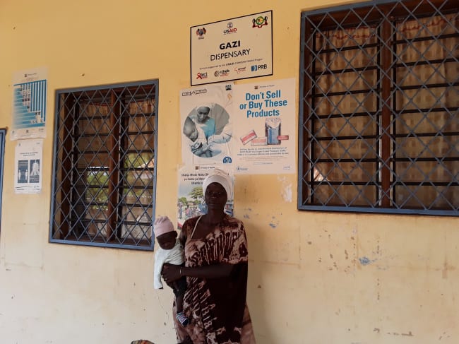 Young mother standing holding a baby at Gazi dispensary, with white head wrap. Saumu Bakari, from a neighboring Zero Zero village that is yet to have its own dispensary, receives maternal and child health services at Gazi dispensary. The Mikoko Pamoja project continues to support the dispensary.