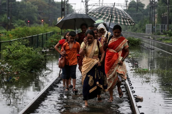 Commuters walk on waterlogged railway tracks after getting off a stalled train during heavy monsoon rains in Mumbai, India, on July 2, 2019. Several women and at least one boy are walking in a group. One of them is talking on a cell phone. Several are sharing umbrellas. The water, even on the train tracks, which are presumably slightly elevated, is up to their ankles. REUTERS/Francis Mascarenhas
