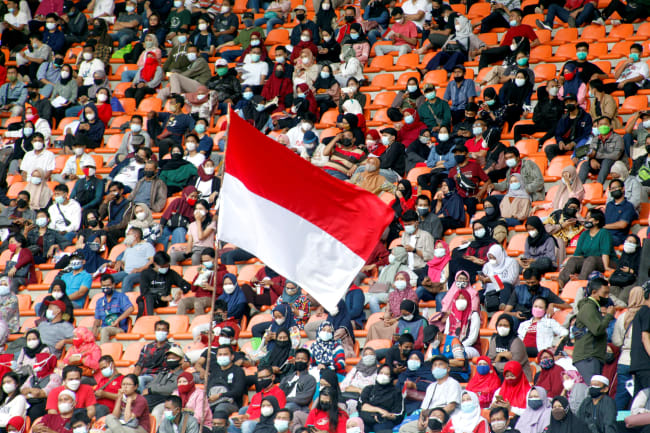 The Indonesian flag flies as people, wearing protective face coverings, wait to receive a COVID-19 vaccine dose at Pakansari Stadium, in Bogor, Indonesia, on August 14, 2021.