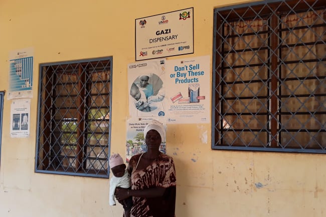 Young mother standing holding a baby at Gazi dispensary, with white head wrap. Saumu Bakari, from a neighboring Zero Zero village that is yet to have its own dispensary, receives maternal and child health services at Gazi dispensary. The Mikoko Pamoja project continues to support the dispensary.