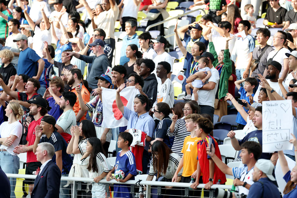 Fans stand in the stadium watching the Japan team train for the World Cup, raising arms and Japanese flags in support.