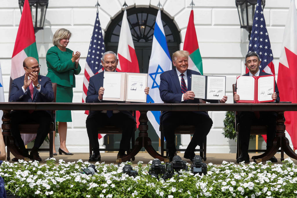Bahraini Foreign Minister Abdullatif Al Zayani, Israeli Prime Minister Benjamin Netanyahu, U.S. President Donald Trump, and Emirati Foreign Minister Abdullah bin Zayed sit at a long table on the White House lawn during the Abraham Accords signing ceremony. Netanyahu, Trump, and bin Zayed hold open folders displaying the signed documents. Emirati, U.S., Bahraini, and Israeli flags line the portico behind them, and a woman in a green suit applauds at left.