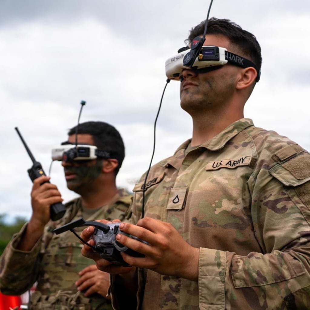 Two soldiers hold controllers and wear tech head pieces as they operate a first-person-view drone during a live-fire exercise.