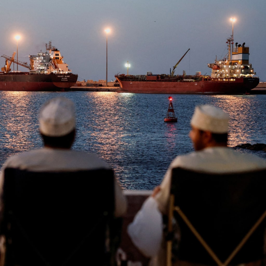 Two men sit facing away, looking out toward two ships docked in the water.