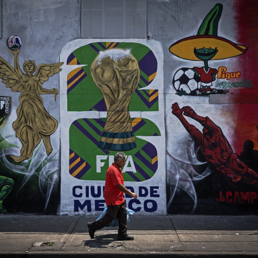 A man walks down a sidewalk in front of a concrete wall displaying a painted mural for the 2026 World Cup in Mexico.