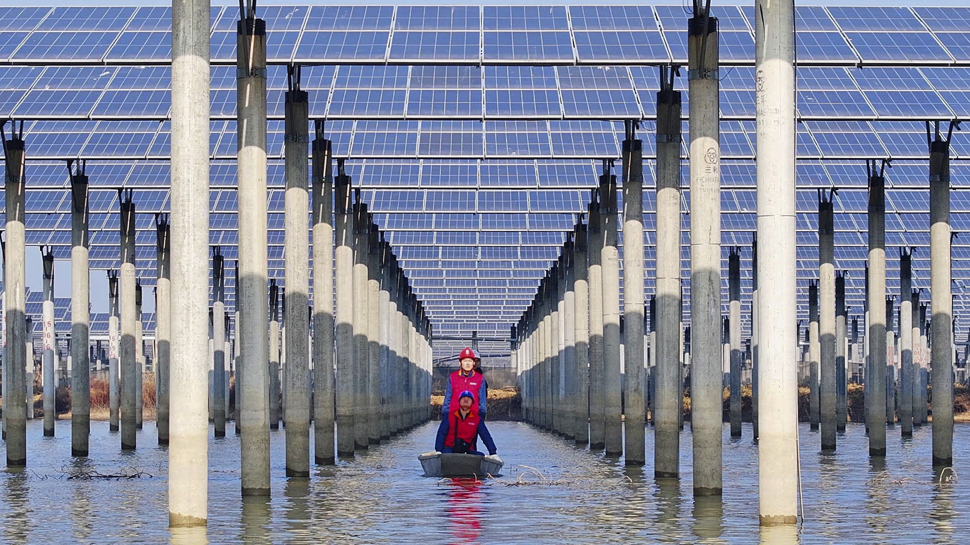 Workers check solar panels installed over a lake from underneath while in a boat.