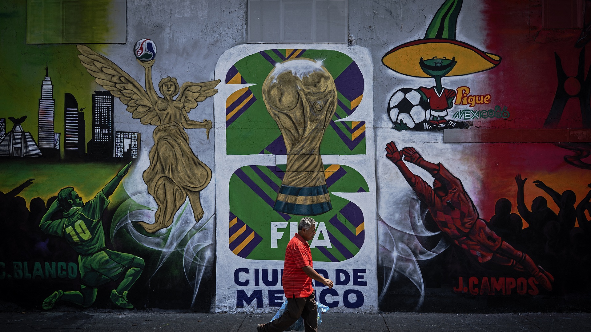 A man walks down a sidewalk in front of a concrete wall displaying a painted mural for the 2026 World Cup in Mexico.
