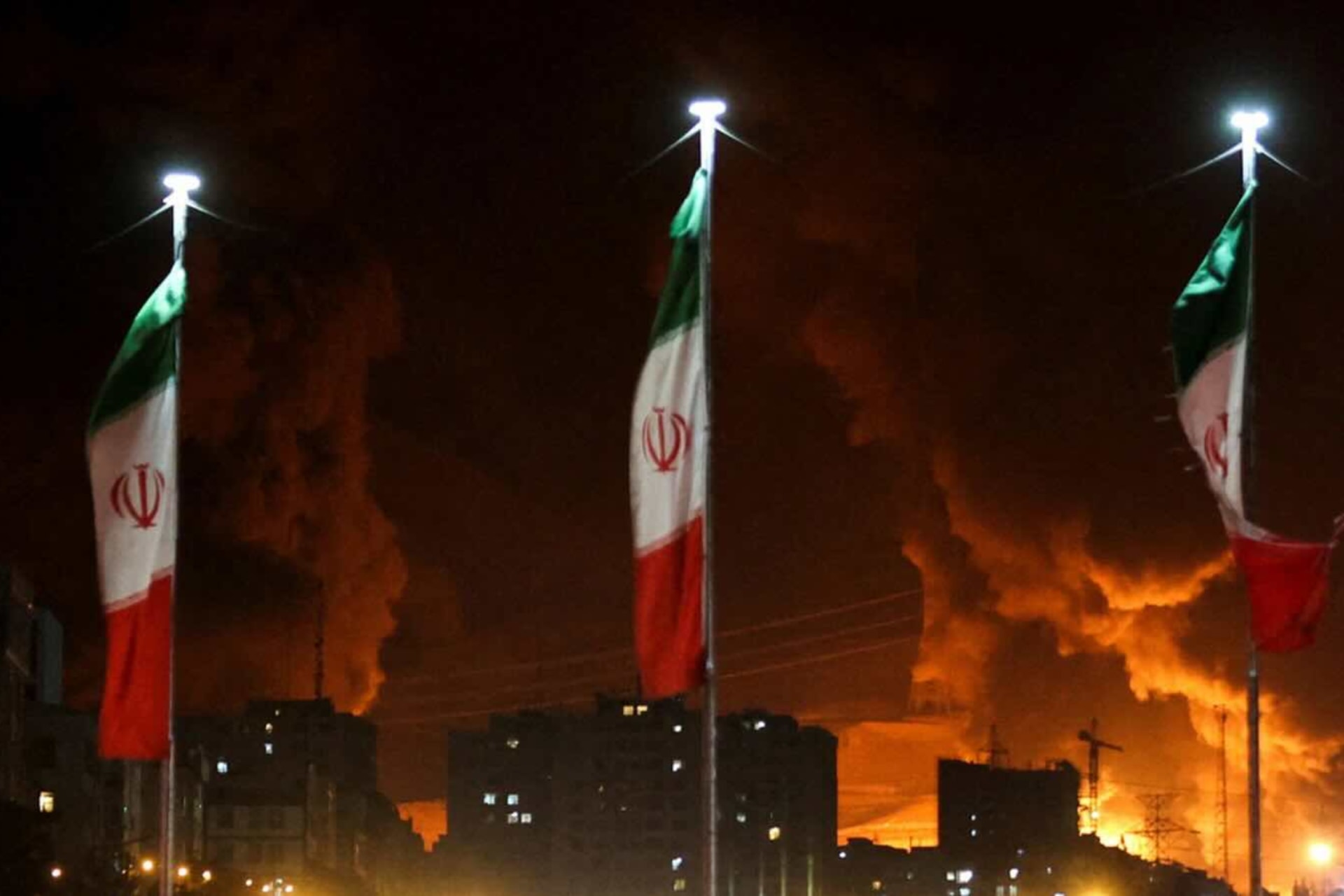 <p>Iranian flags fly as fire and smoke from an Israeli attack on Sharan Oil depot rise, following Israeli strikes in Tehran on June 15, 2025.</p>