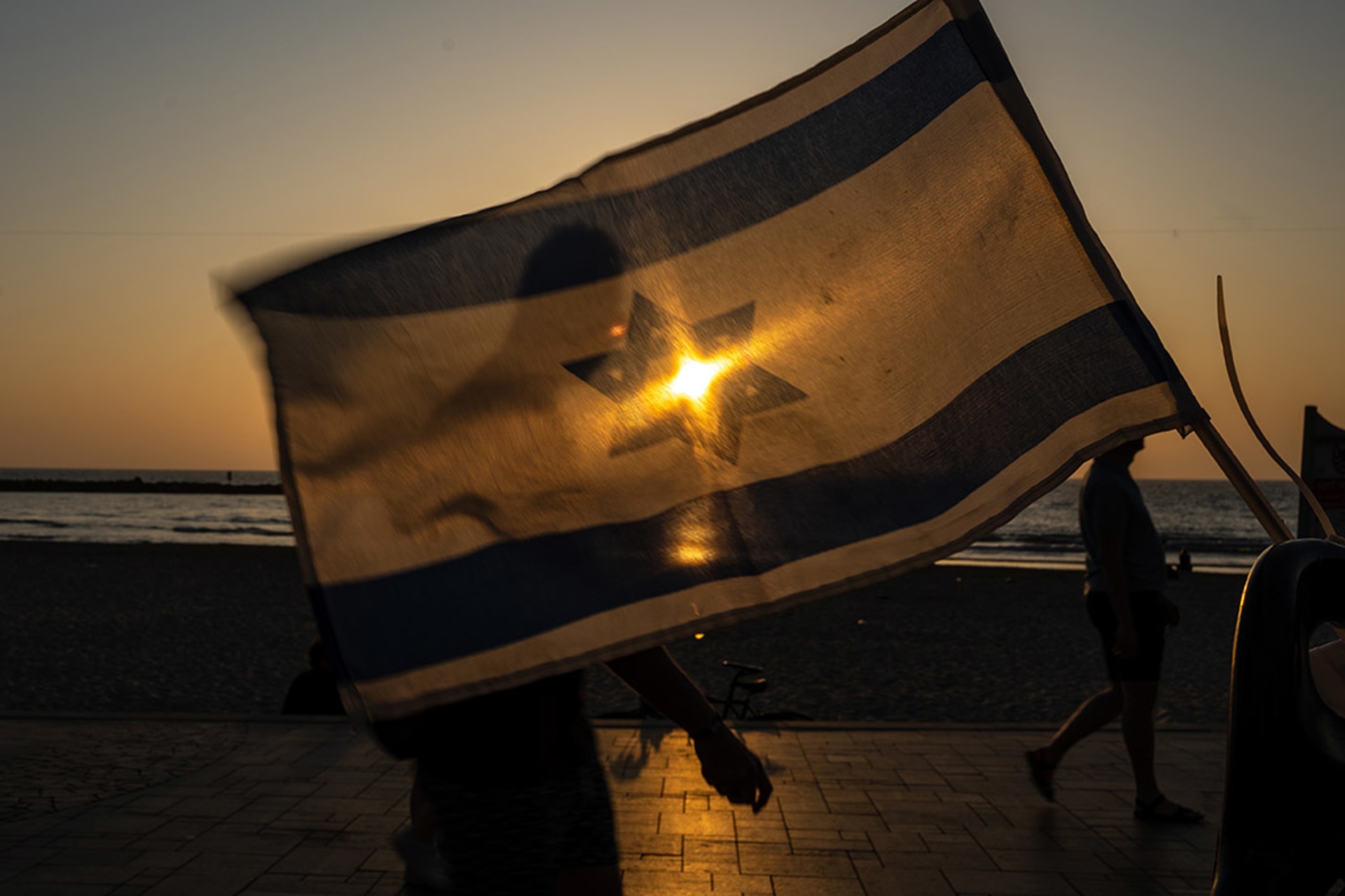 <p>People walk past a waving Israeli flag as the sun on May 25, 2023 in Tel Aviv, Israel.</p>