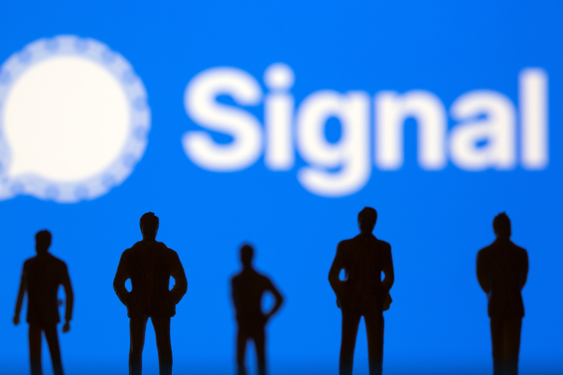 <p>Small toy figures are seen in front of Signal logo.</p>

