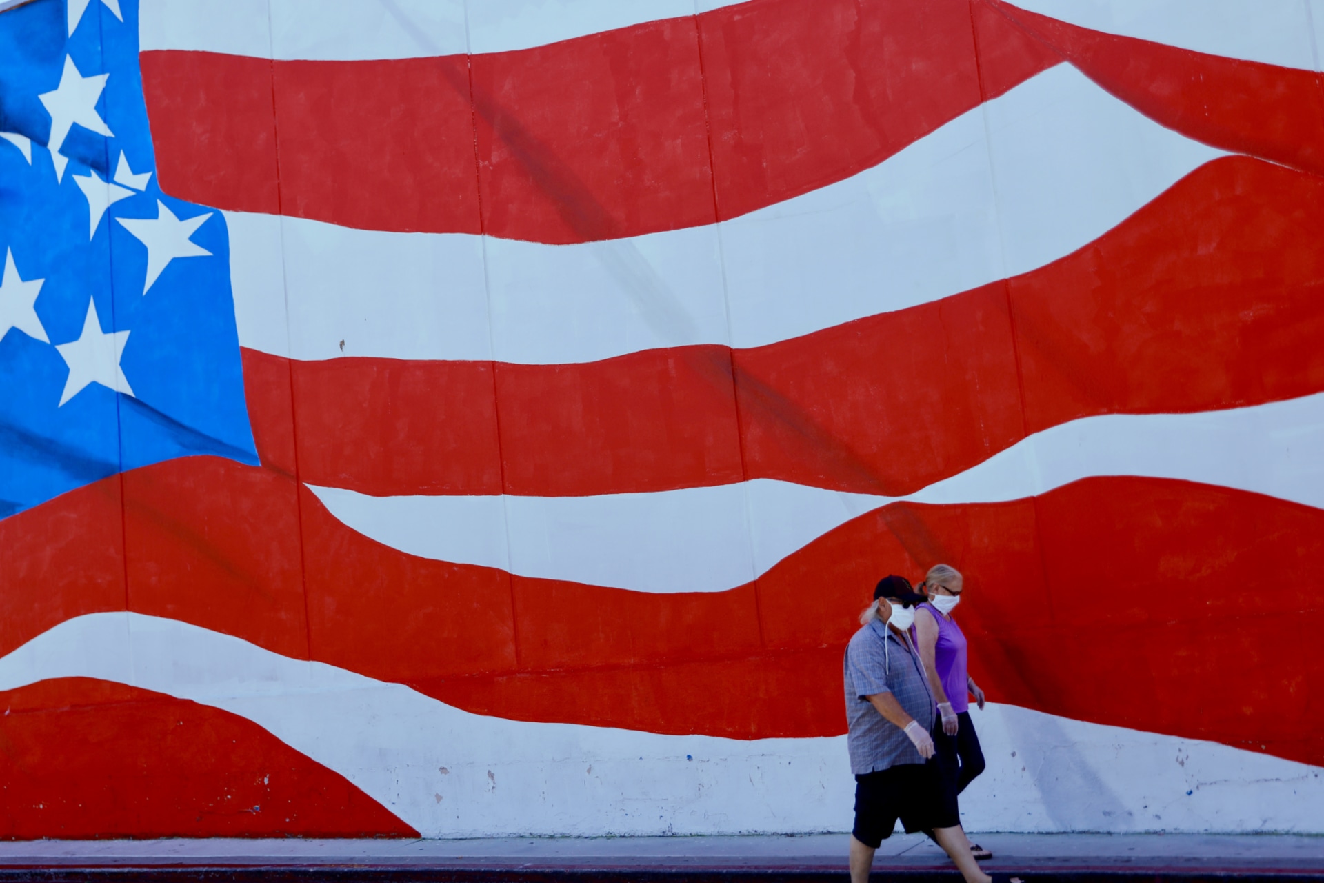 <p>Two people in masks walk by a mural of the U.S. flag in Ocean Beach, California.</p>

