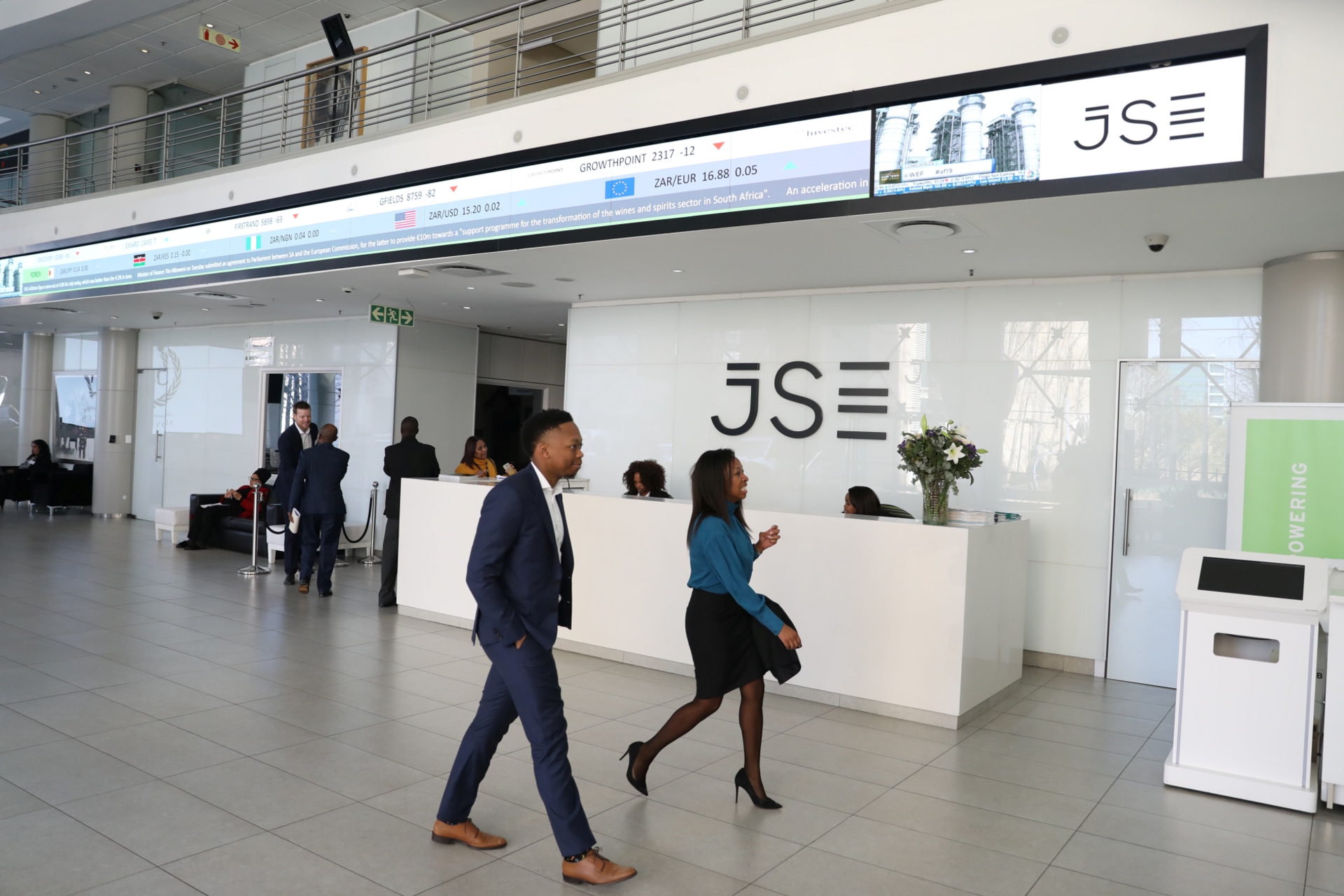 <p>Visitors walk past a reception with an electronic board displaying movements in major indices at the Johannesburg Stock Exchange building in Sandton, Johannesburg, South Africa, August 22, 2019.</p>
