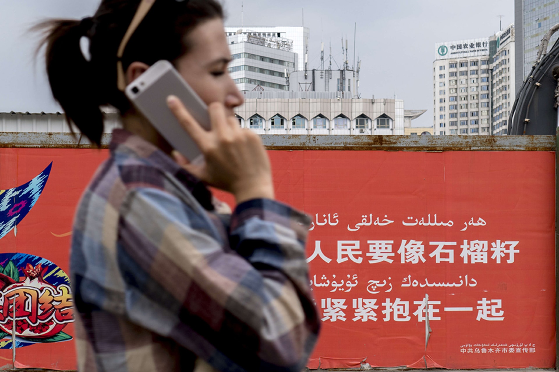 <p>A young Uygur girl makes a phone call with her iPhone while walking past a propaganda wall.</p>
