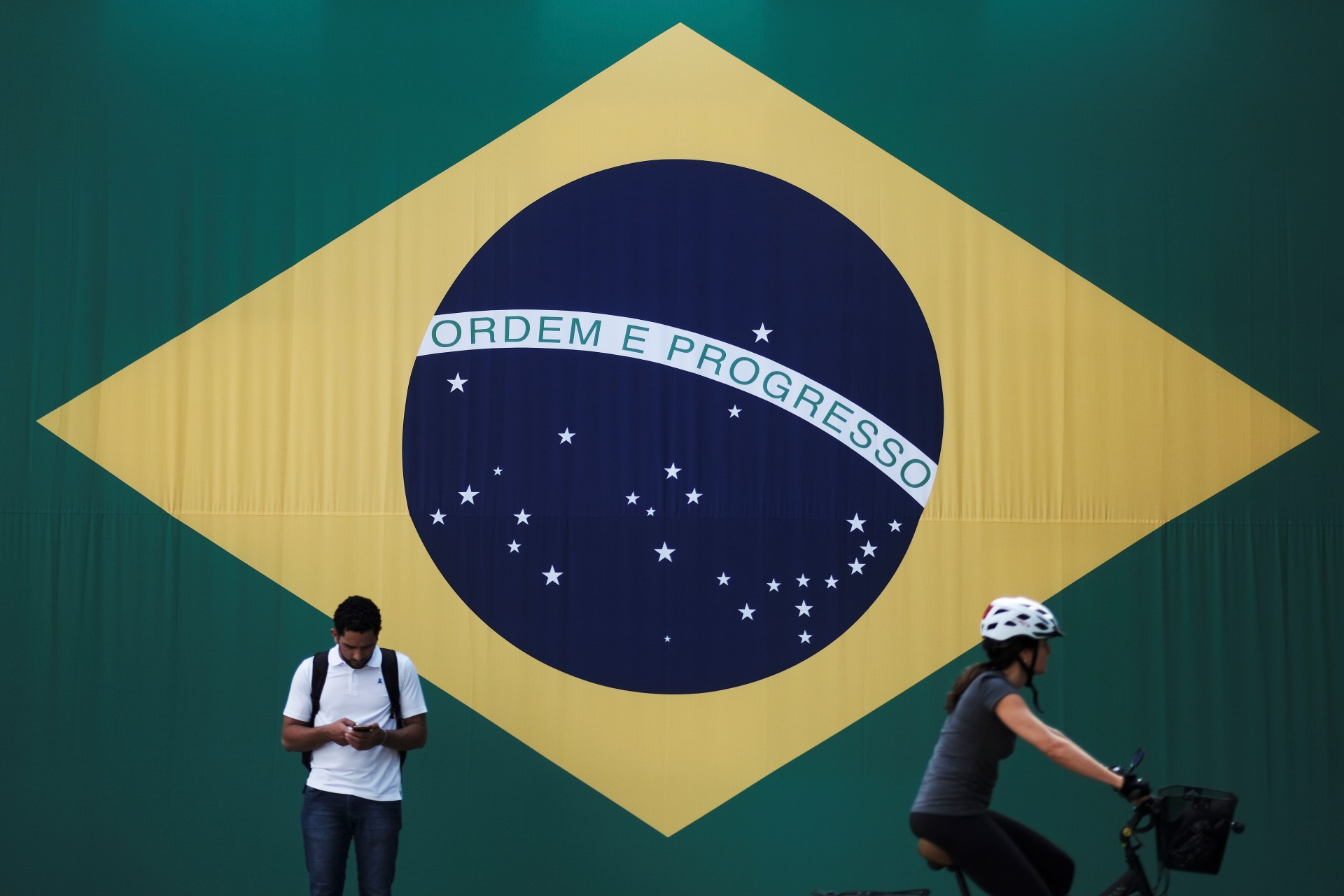 <p>A man checks his mobile phone as a woman riding a bike passes next to a big Brazilian flag in Sao Paulo, Brazil June 28, 2018. </p>
