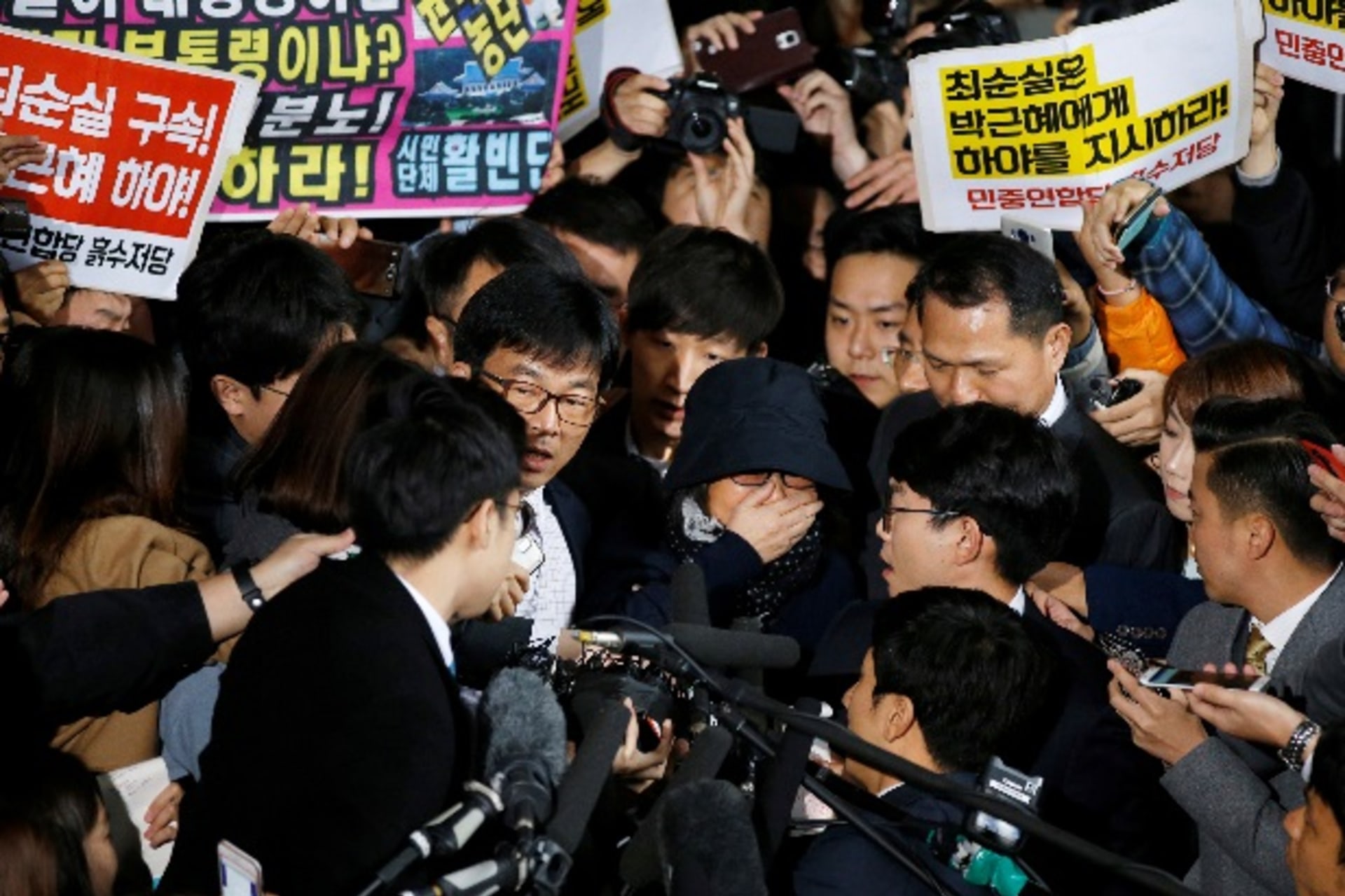 <p>Choi Soon-sil (C), who is involved a political scandal, reacts as she is surrounded by media and protesters upon her arrival a…a, October 31, 2016. The banner (top L) reads “Arrest Choi Soon-sil, Call for Park Geun-hye to step down”. REUTERS/Kim Hong-Ji</p>
