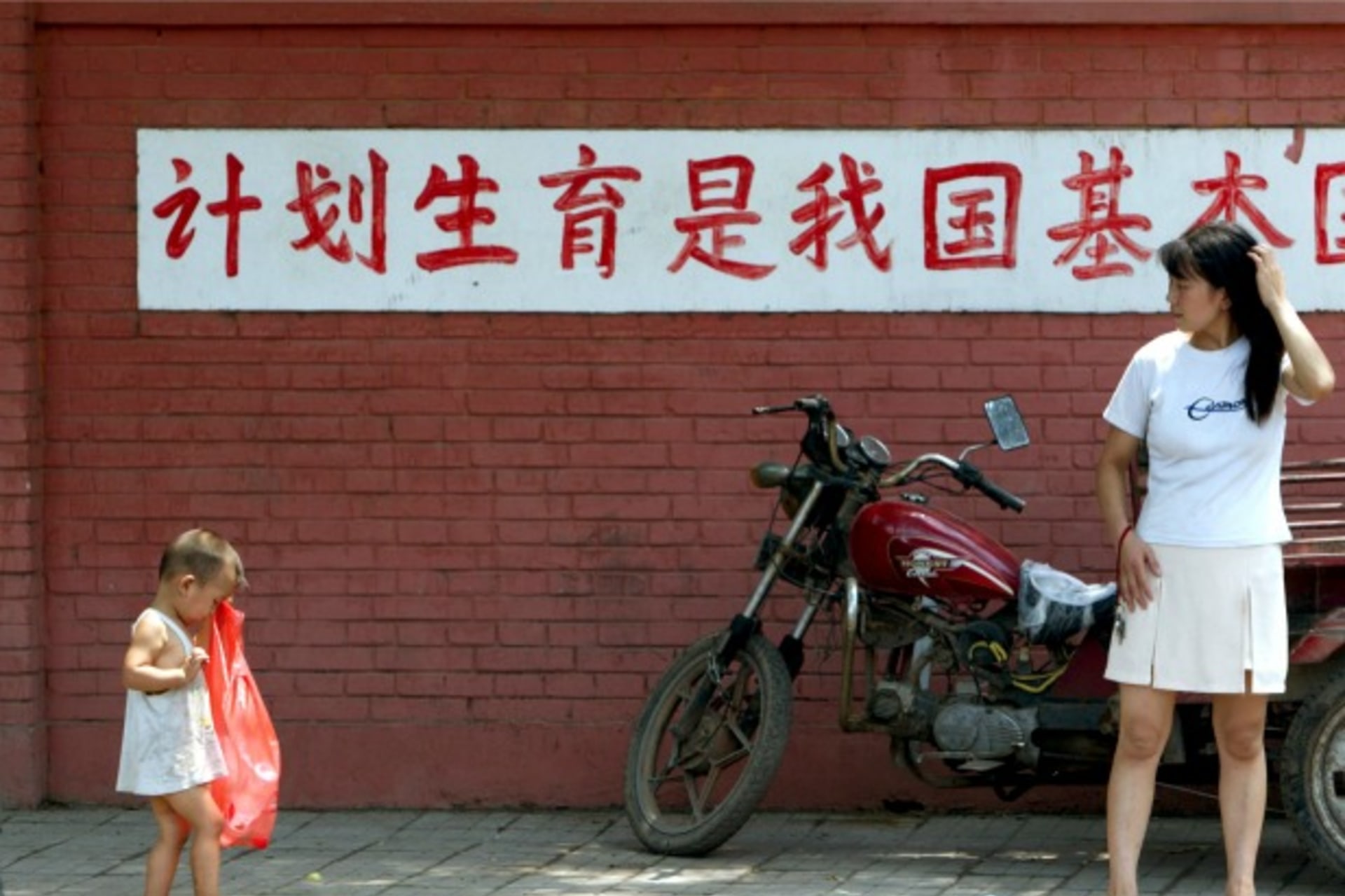 <p>A young Chinese mother watches her child in front of a sign reading “birth control is a basic state policy of our country” in Beijing on July 23, 2002.</p>
