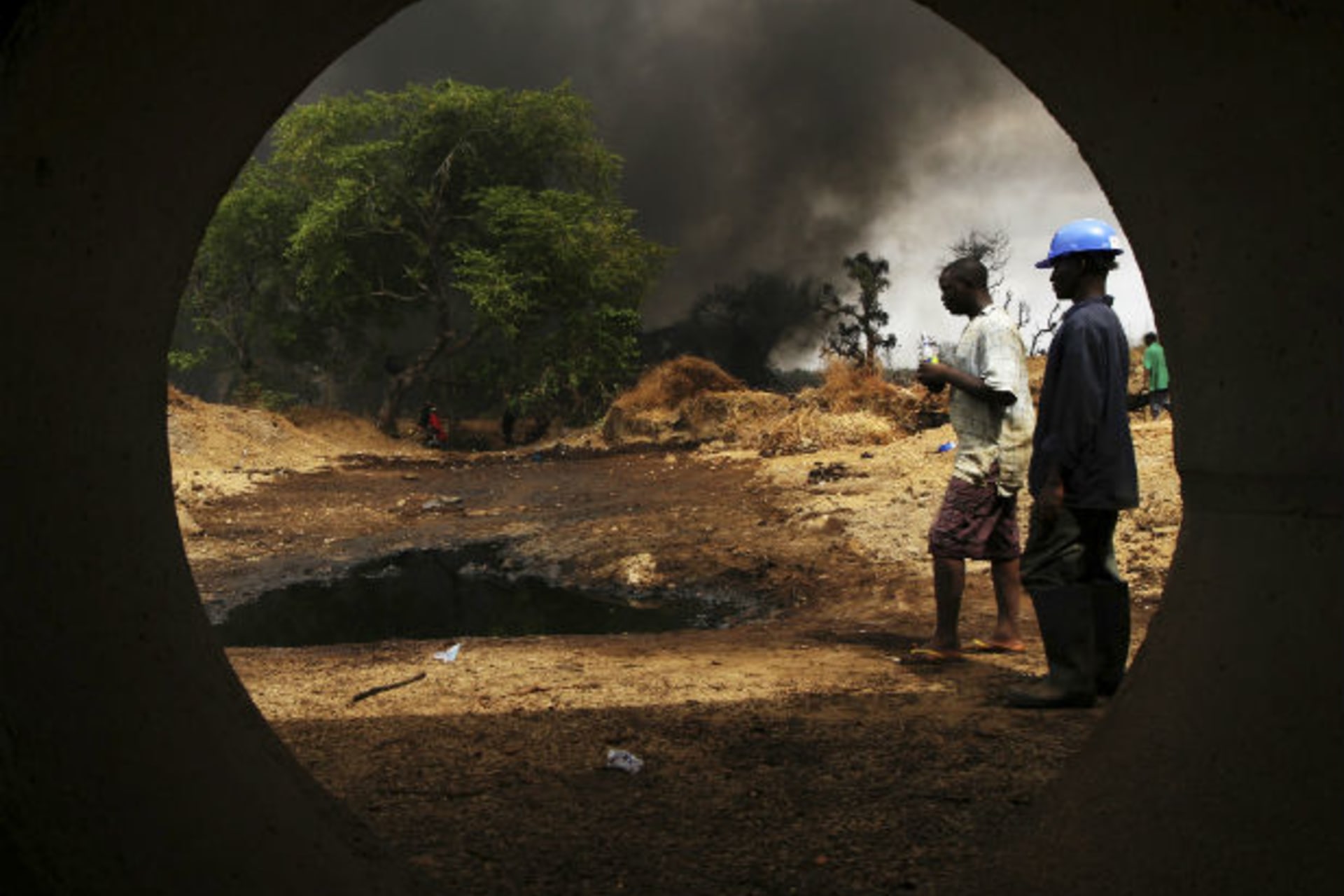 Workers, seen through a pipe, look on at the scene of an oil pipeline fire in Dadabili, Niger state 02/04/2011.