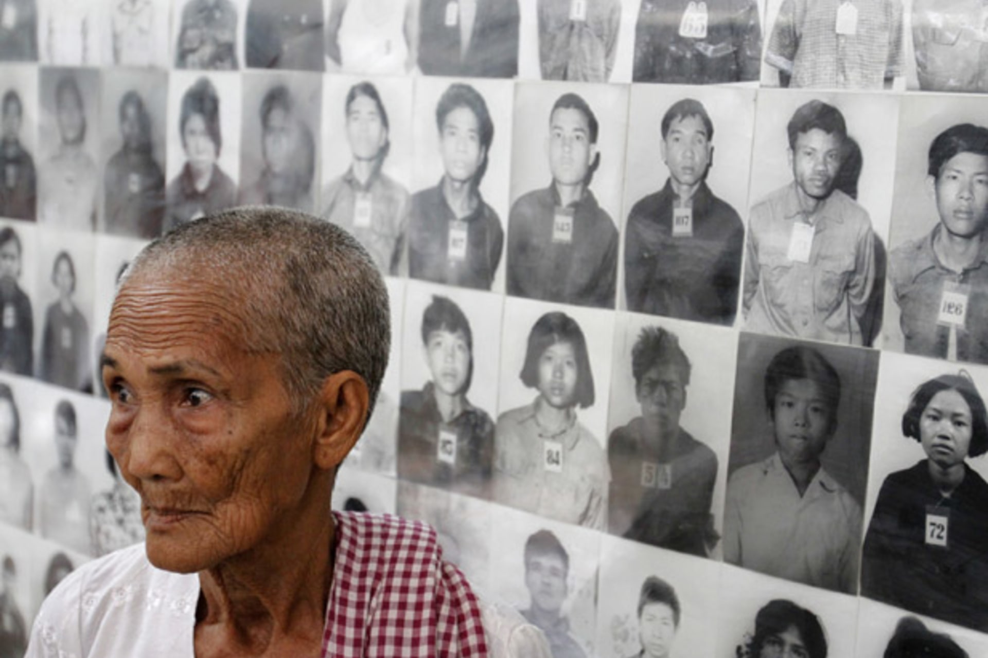 A survivor of the Khmer Rouge regime Hem Sakou stands in front of portraits of victims at the Tuol Sleng (S-21) genocide museum in Phnom Penh
