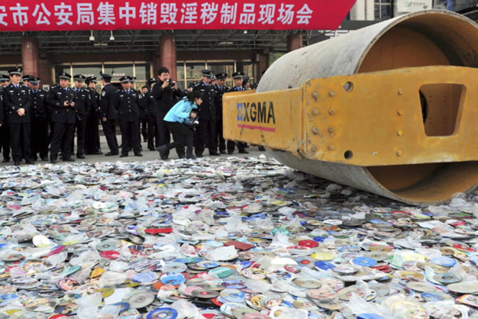 Police officers look on as a road roller is used to destroy confiscated pornographic DVDs and pirated publications in Xi'an
