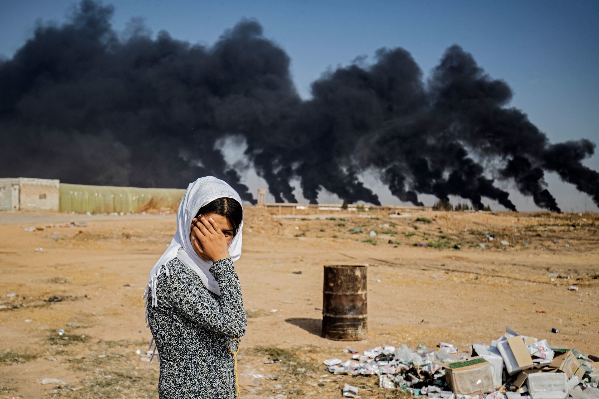 <p>A woman covers her face as she stands on the outskirts of a town near the Syria-Turkey border, while smoke plumes meant to decrease visibility for Turkish warplanes billow from tire fires during a cross-border offensive, on October 16, 2019.</p>
