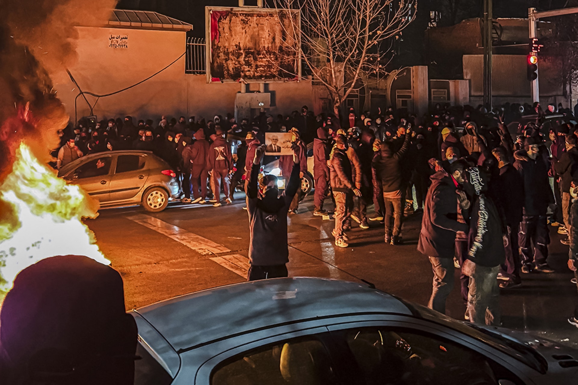 A large crow of Iranians gather around cars and buildings, blocking a street as a fire billows up on the left during protests in Tehran.