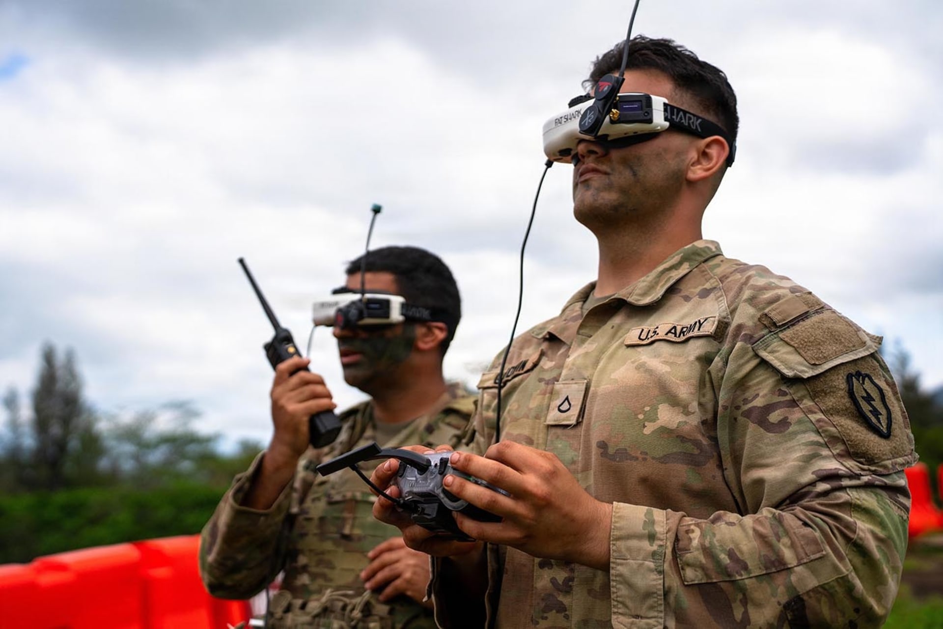 Two soldiers hold controllers and wear tech head pieces as they operate a first-person-view drone during a live-fire exercise.