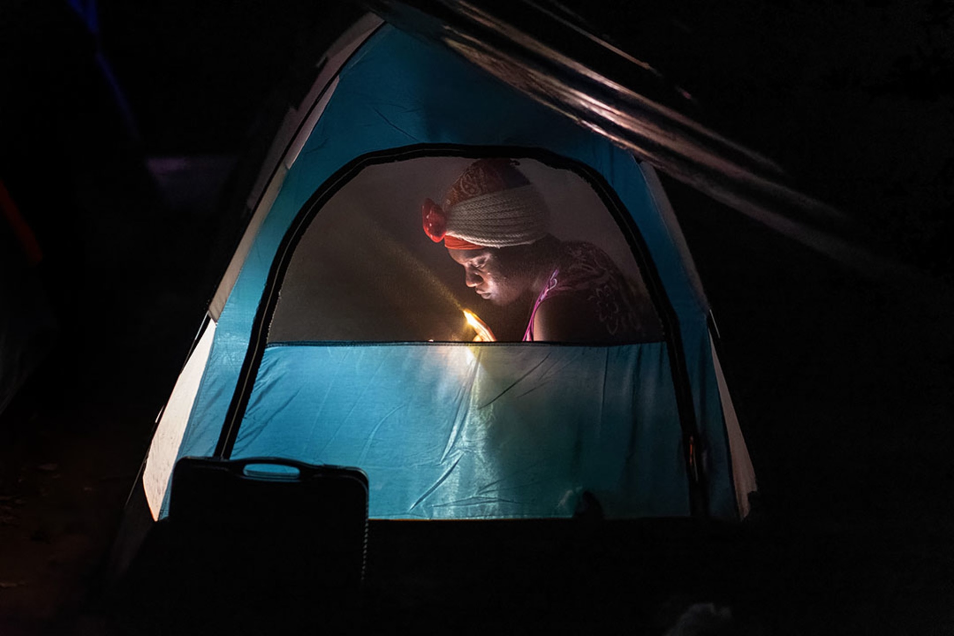 A woman immigrant from Haiti is seen through the screen of a tent reading by flashlight.
