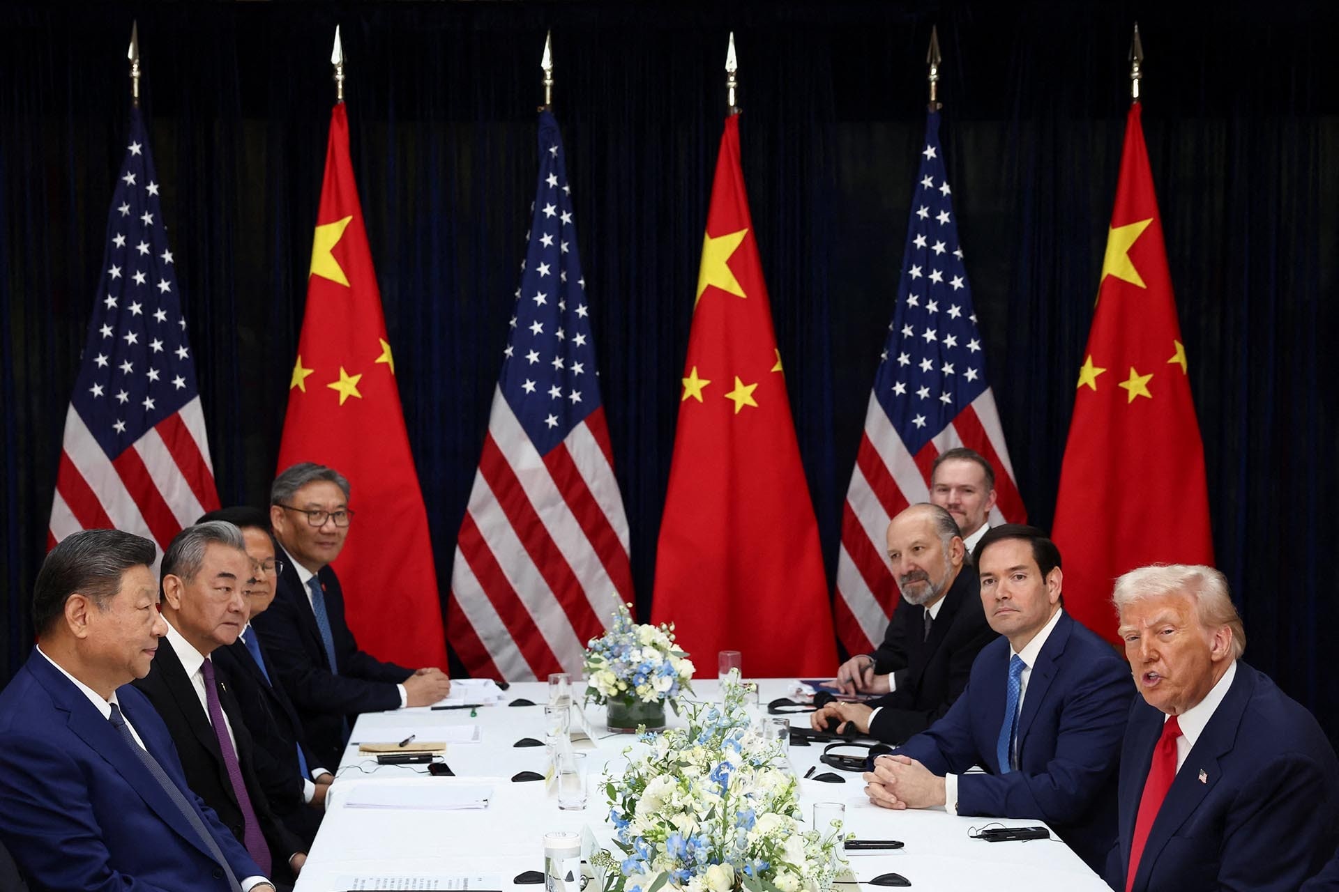 President Trump and President Xi sit across from the table from each other. Other men sit beside them. Chinese and American flags stand in the background