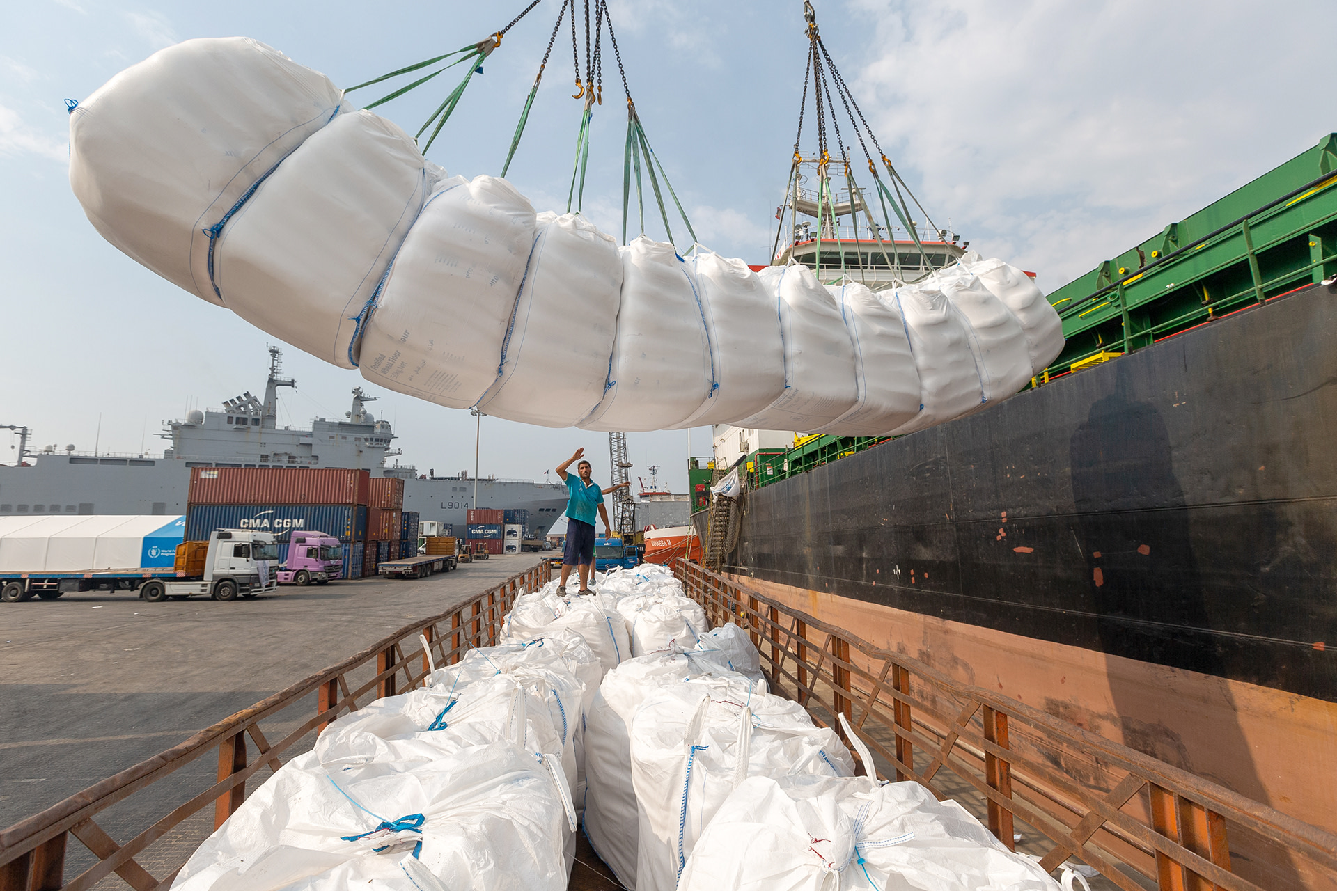 A crane lowers a white package containing food aid into a crate off of the side of a ship.
