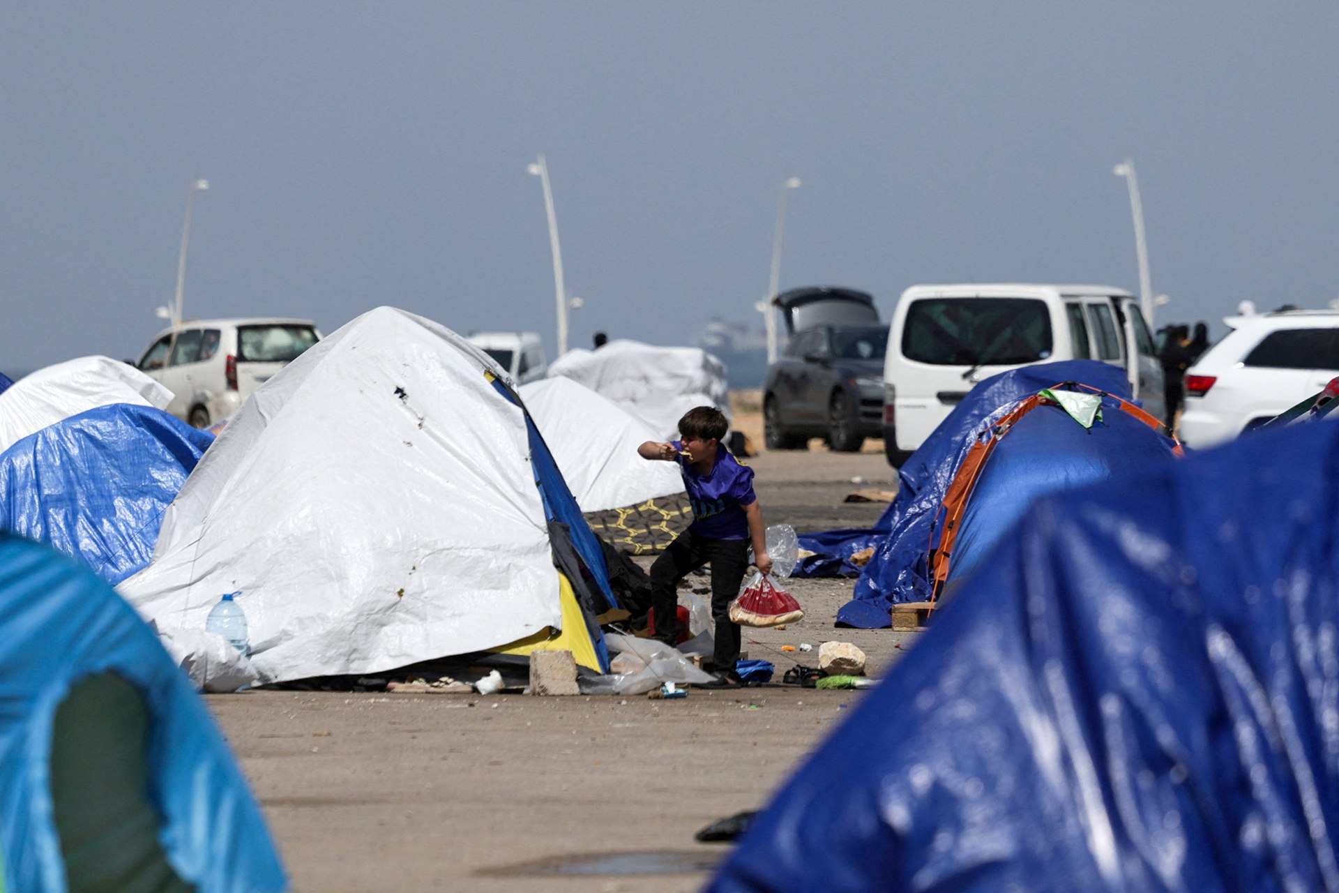 A child stands outside a tent, eating food, surrounded by other tents and vehicles on open land.