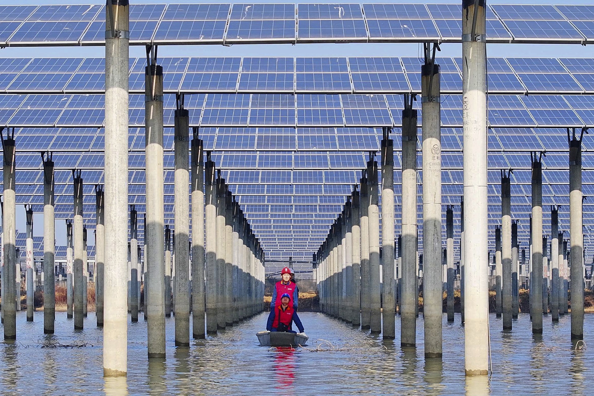 Workers check solar panels installed over a lake from underneath while in a boat.