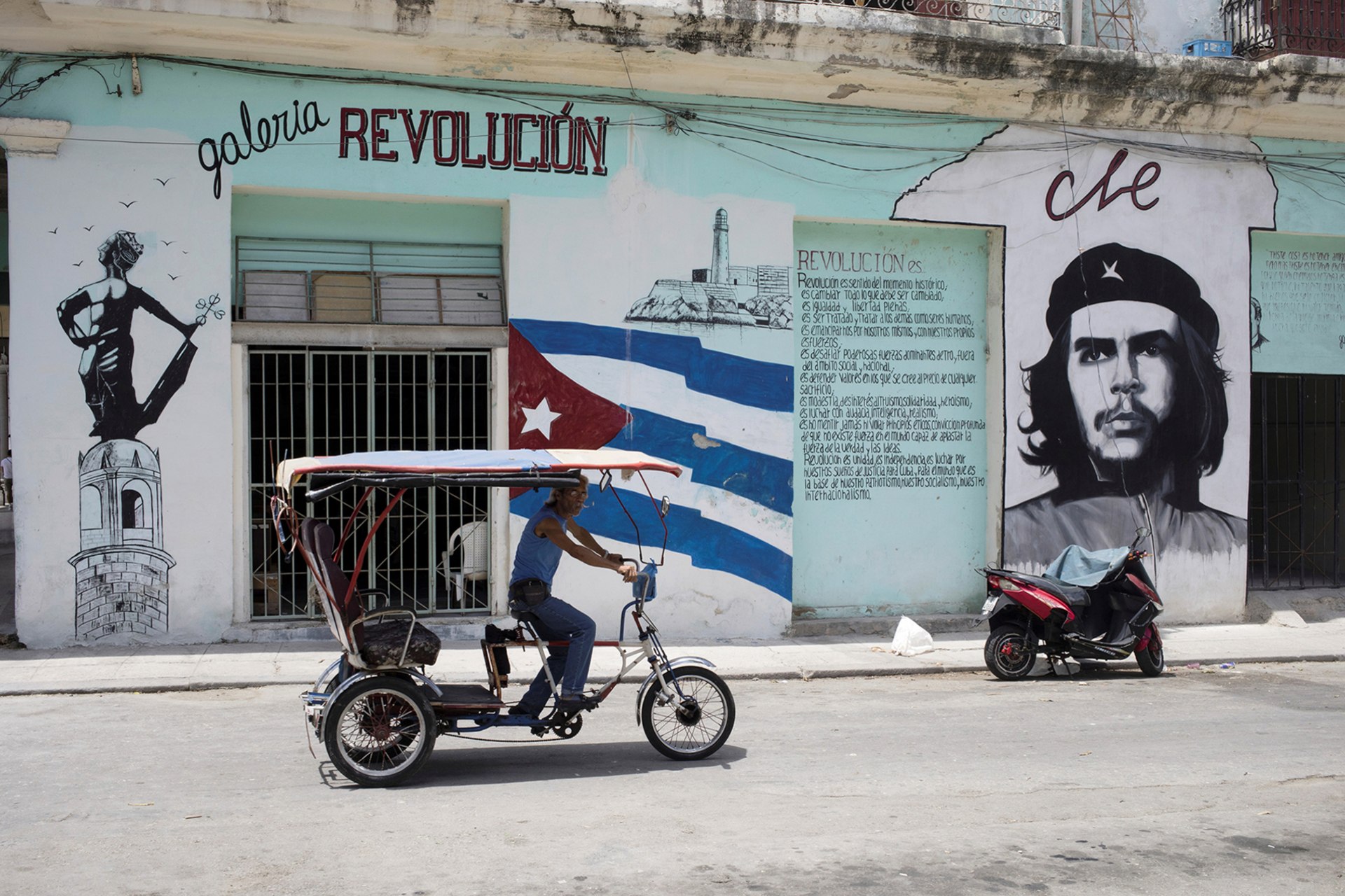 A bicycle taxi passes in front of a building decorated with murals to the glory of the revolution and Che in the old town of Havana.