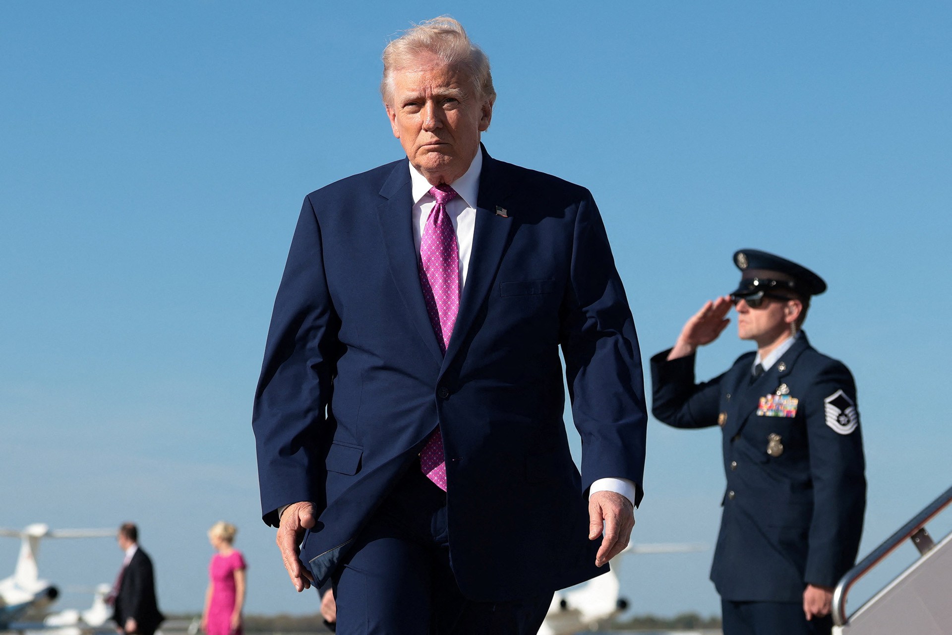 U.S. President Donald Trump walks to board Air Force One on his way to Virginia, at Joint Base Andrews in Maryland, on April 10, 2026. An officer in a blue uniform salutes behind him.