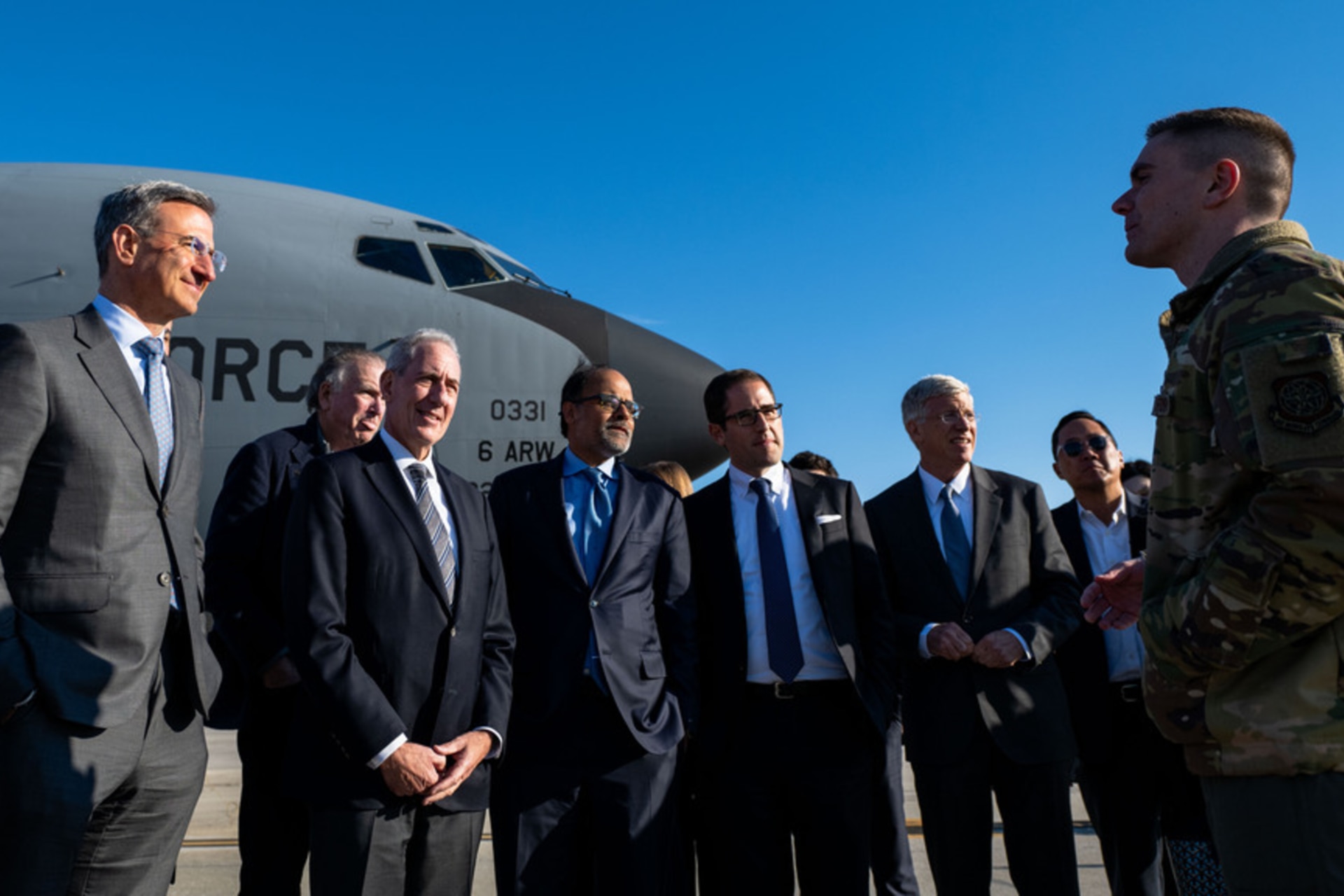 A group of men in suits stand in front of a grey military aircraft.
