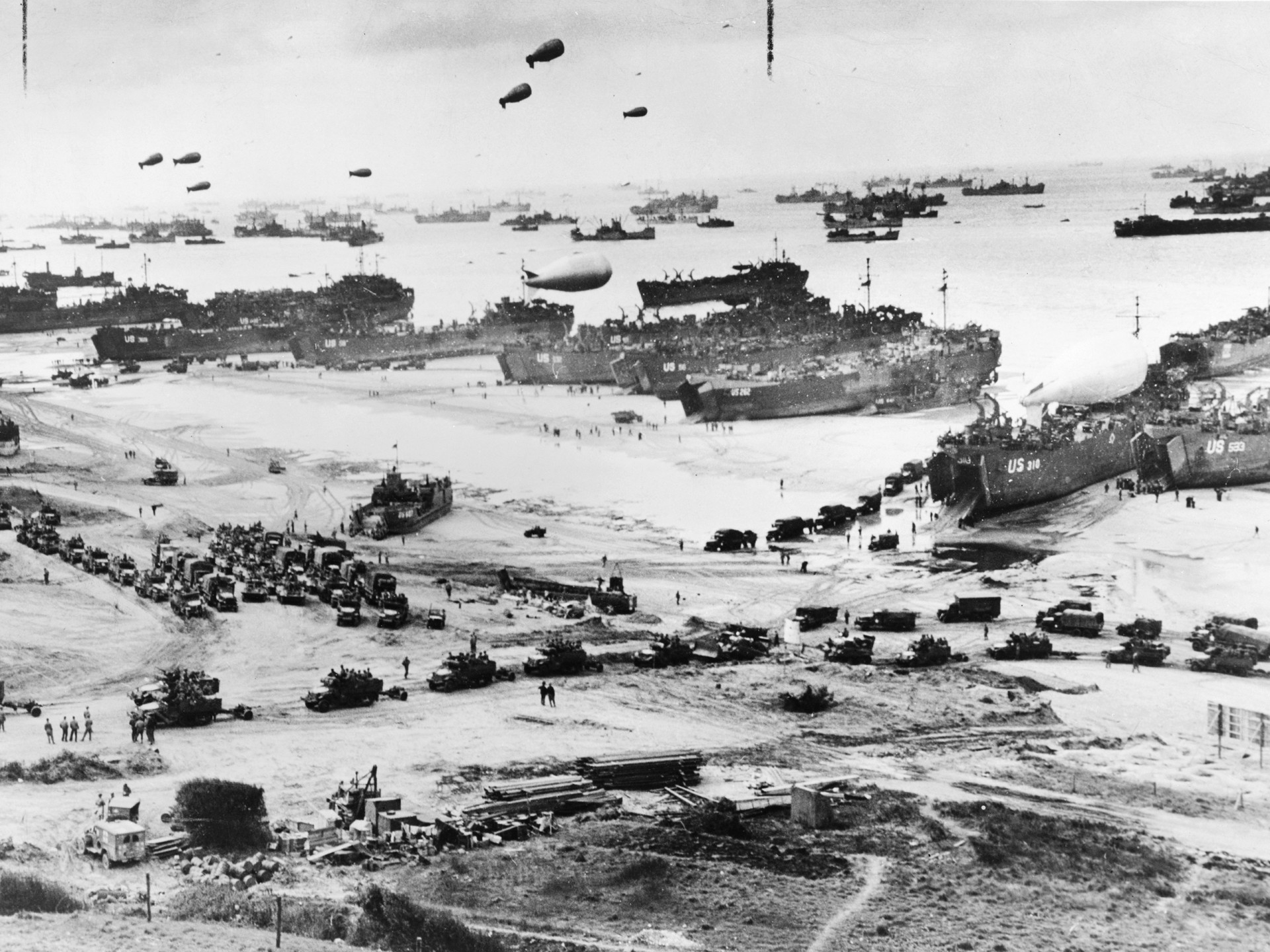Bird's-eye view of landing craft, barrage balloons, and allied troops landing in Normandy, France on D-Day.